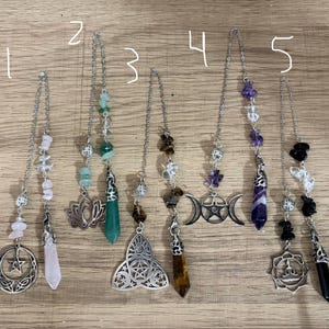 Pode incluir: Cinco marcadores de página de metal prateado, cada um com um pingente pendurado e uma ponta de pedra preciosa. Os pingentes incluem uma lua crescente com estrela, uma flor de lótus, uma triquetra, uma lua tripla com pentáculo e um mandala.