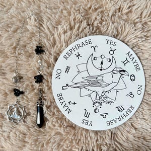 May include: A divination set comprising a pendulum with black beads and a silver charm, and a white circular board. The board features a raven illustration, zodiac symbols, and the words "Yes", "No", "Maybe", and "Rephrase".