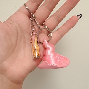 Op de afbeelding: Een sleutelhanger met een klein doorzichtig glazen flesje gevuld met gedroogde gele bloemblaadjes, afgewerkt met roze druipende hars, en een aparte bedel in de vorm van een heksenhoed van roze hars aan een zilverkleurige metalen ring.