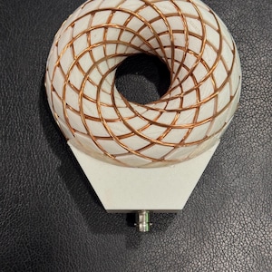 May include: A white, donut-shaped sculpture with a copper wire grid design. The sculpture sits atop a white, geometric base with a silver connector. The object is displayed on a black, textured surface.