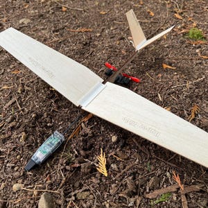 May include: A wooden glider airplane with a white finish and black string. The wings are labeled "Free Flight" and "Free Flight".