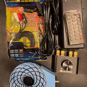 Könnte beinhalten: Eine Ansammlung elektronischer Komponenten, darunter ein schwarzer Verstärker mit goldfarbenen Eingängen, eine Fernbedienung und verschiedene Kabel. Ein blaues und kupferfarbenes kugelförmiges Objekt ist ebenfalls vorhanden, zusammen mit einem Netzteil.