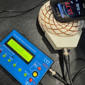 Op de afbeelding: Een blauwe FG-200 DDS-functiesignaalgenerator met een digitaal display met golf- en frequentiemetingen. Een zwart apparaat geeft een magnetische veldmeting van 33,77 μT weer. Een witte en koperdraadbol is aangesloten op de apparaten.