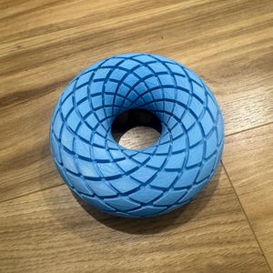 May include: A light blue, 3D-printed torus with a geometric pattern. The design features a series of intersecting lines creating a textured surface. The object is displayed on a wooden surface.