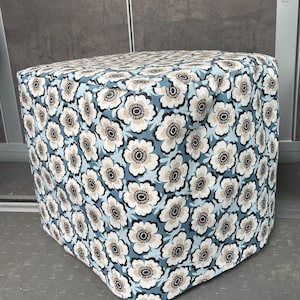Può includere: Un pouf quadrato rivestito in tessuto floreale. Il tessuto presenta un motivo di fiori bianchi, beige e neri su sfondo azzurro. Il pouf ha una forma solida, a forma di cubo.