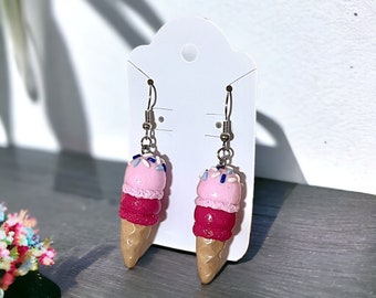 May include: A pair of dangle earrings featuring a pink, red, and brown ice cream cone design with white and blue sprinkles.