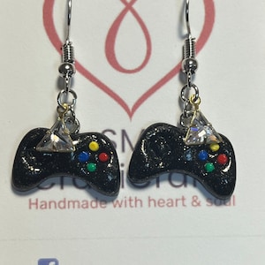 May include: Black glitter game controller earrings with a clear rhinestone on top. The earrings have silver hooks.