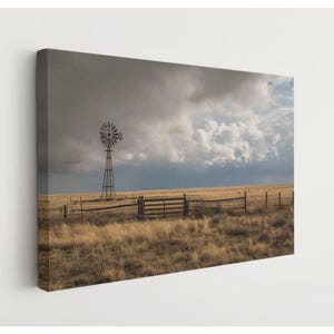 May include: A canvas print of a rural landscape featuring a windmill, wooden fence, and tall grass under a dramatic, cloudy sky. The color palette is dominated by muted browns, yellows, and grays, creating a rustic and serene atmosphere.