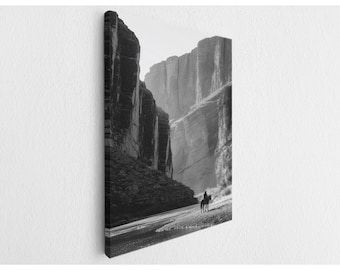 May include: A black and white canvas print featuring a landscape of towering canyon walls flanking a river. A lone figure on horseback stands on the riverbank in the foreground, set against the vast, rugged rock formations of the deep canyon.