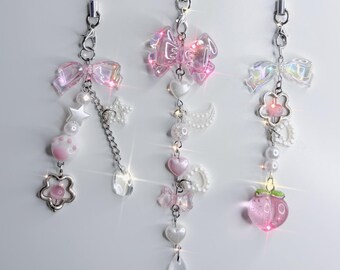 Puede incluir: Tres charms para teléfono de color rosa e iridiscente con detalles de perlas y cristales. Cada charm presenta un lazo rosa, un corazón, una estrella, una flor, una luna creciente y un melocotón.