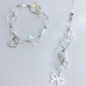 Op de afbeelding: Een witte armband- en telefoonbedelset. De armband heeft iriserende vlinder- en bloembedels, witte parels en een zilveren wolkenbedel. De telefoonbedel heeft witte kralen, een strik en een varkensbedel.