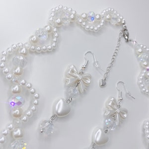 A white pearl twirl necklace with iridescent beads and pearl bows with in the middle matching earrings with a pearl bow, iridescent beads and pearl hearts.