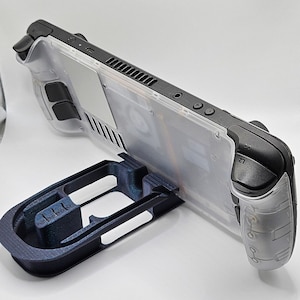 May include: A clear plastic handheld gaming console with black buttons and a black stand. The console has a clear plastic back panel with a black vent and a black trim around the edges.