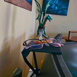 May include: A black metal monitor stand with a black mesh surface. Two pairs of glasses and two cookies are on the stand. A green dragon figurine is in the background.