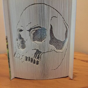 Könnte beinhalten: Ein weißes Buch, dessen Seiten gefaltet sind, um eine 3D-Schädelskulptur zu erstellen.