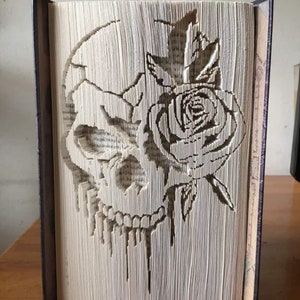 May include: A book with a skull and rose design created by folding the pages. The skull is on the left side of the book and the rose is on the right side. The design is in black and white.