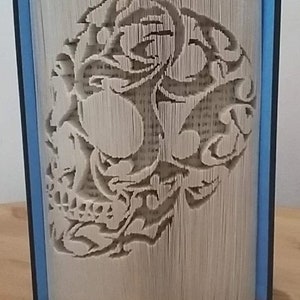 Op de afbeelding: Een boeksculptuur met een schedelontwerp uitgesneden in de pagina's. De randen van het boek zijn blauw, de pagina's beige. Het schedelontwerp is gedetailleerd en toont de vaardigheid van de kunstenaar.