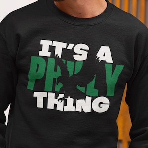 May include: Black crew neck sweatshirt with the text "IT'S A PHILLY THING" in white and green. The word "PHILLY" features a silhouette of a bird. A casual, comfortable top.
