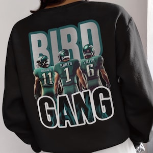以下が含まれることがあります： 黒いスウェットシャツ。緑色のユニフォームを着た3人のアメリカンフットボール選手のグラフィック。選手の上には「BIRD」、下には「GANG」の文字。選手の名前と背番号は、ブラウン11、ハーツ1、スミス6。