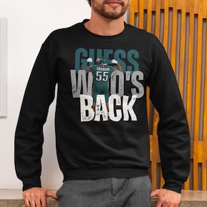 以下が含まれることがあります： 白とティール色の「GUESS WHO'S BACK」という文字が入った黒いスウェットシャツ。デザインは、名前「GRAHAM」と番号「55」が入った緑色のユニフォームを着たフットボール選手が特徴です。