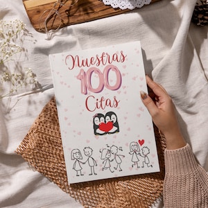 Könnte beinhalten: Ein weißes Journal mit dem Text "Nuestras 100 Citas" in rosa und lila Schrift. Das Journal zeigt ein Cartoon-Bild von zwei Pinguinen, die ein rotes Herz halten. Es gibt auch Cartoon-Bilder von Paaren in verschiedenen Posen.