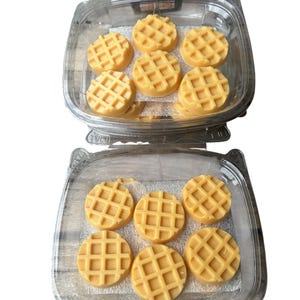 Op de afbeelding: Twee doorzichtige plastic containers gevuld met ronde, lichtgele wafels. Elke wafel heeft een rasterpatroon. De wafels zijn in twee lagen gerangschikt, waarbij de bovenste container acht wafels en de onderste container zeven wafels bevat.