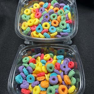 May include: Two clear plastic containers filled with colourful cereal rings. The cereal is a mix of yellow, orange, red, blue, green, and purple.