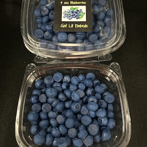 May include: Two clear plastic containers filled with blue wax melts shaped like blueberries. One container has a label that reads "4 ozs Blueberries" and "Get Lit Embeds" with a blueberry graphic.
