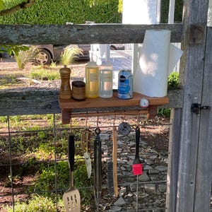 Peut inclure: Une étagère murale en bois avec une tige horizontale munie de cinq crochets en cuivre. L'étagère supporte un moulin à poivre en bois, deux flacons souples transparents, une canette d'eau gazeuse bleue et un rouleau d'essuie-tout. Des ustensiles sont suspendus, dont une spatule en métal, des pinces, une brosse et un pinceau à pâtisserie en silicone rouge.