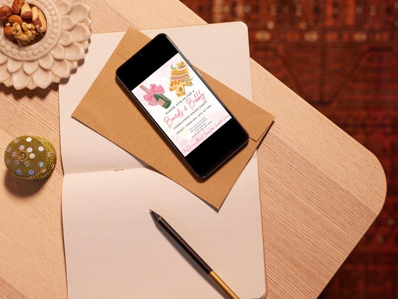 May include: A smartphone displaying a pink and gold invitation for a "Bride & Bubbly" party. The invitation features a champagne bottle and a cake with pink and gold decorations. The text on the invitation reads "Please join us for a Bride & Bubbly Charcuterie Board Party. Friday February 10th at 7pm.  RSVP to [email protected]"