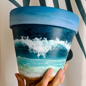 May include: A terracotta flower pot painted with a blue and white ocean scene. The pot features a blue sky, white foamy waves, and a turquoise ocean.