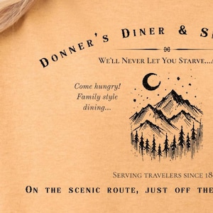 May include: A tan t-shirt with a black graphic design that reads "Donner's Diner & Snow Lodge" with a mountain scene and the text "We'll never let you starve...again."  The graphic also includes the text "Come hungry! Family style dining..." and "Serving travelers since 1846."  The bottom of the graphic reads "On the scenic route, just off the California Trail."