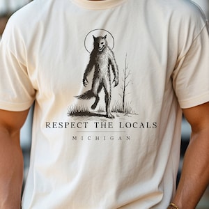 May include: A cream-colored t-shirt with a black and white illustration of a werewolf. The text "Respect the Locals Michigan" is printed below the illustration.