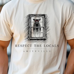 May include: A white t-shirt with a black and white illustration of a window with a creature inside. The text "RESPECT THE LOCALS AMITYVILLE" is printed below the illustration.