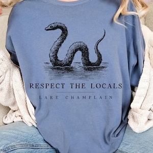 May include: A blue t-shirt with a black graphic of a sea serpent and the text "Respect the Locals Lake Champlain".