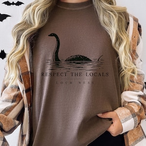 May include: A brown sweatshirt featuring a graphic of the Loch Ness Monster emerging from the water. The text "Respect the Locals" and "Loch Ness" are printed below the illustration in a black font. The sweatshirt has long sleeves.