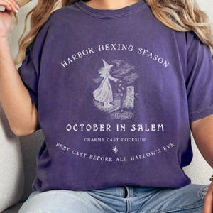 May include: Purple t-shirt with the text "HARBOR HEXING SEASON" and "OCTOBER IN SALEM". The shirt features a white graphic of a witch casting a spell. The text "BEST CAST BEFORE ALL HALLOW'S EVE" is also printed on the shirt.