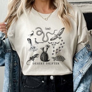 May include: A beige t-shirt with a black and white graphic print of desert animals and plants. The text "DESERT DRIFTER" is printed below the graphic.