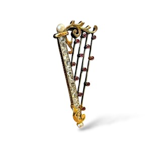 May include: A harp-shaped brooch crafted in gold-tone metal, featuring a vertical row of clear faceted stones, a single white pearl at the top and bottom, and dark metal strings accented with small, round, reddish-pink stones along the frame.