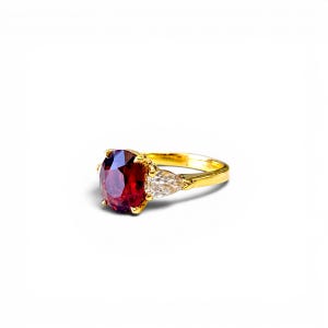 May include: A gold ring featuring a large, oval-cut, deep red gemstone as the primary focal point. Two smaller, pear-shaped, clear gemstones flank the main stone. The ring's band is a polished gold color, and the setting is also gold.