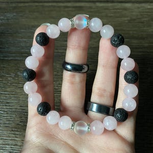 May include: A beaded bracelet featuring alternating light pink and black beads. The bracelet includes clear beads with silver accents. The bracelet is shown on a hand.