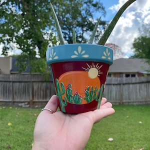 Könnte beinhalten: Ein Terrakotta-Pflanztopf, der mit einer Wüstenlandschaft bemalt ist, mit einem Sonnenuntergang, Kakteen und einer gelben Sonne. Der Topf ist mit weißen Akzenten verziert und enthält eine grüne Sukkulente.