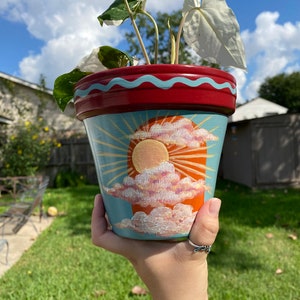 Könnte beinhalten: Ein Terrakotta-Blumentopf mit handbemaltem Design einer Sonne und Wolken. Die Sonne ist orange und gelb, und die Wolken sind rosa und weiß. Der Topf ist in einem blau-roten Farbschema bemalt.