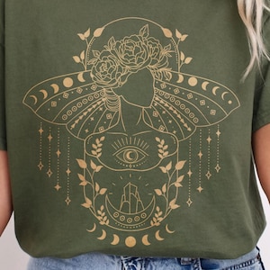 May include: Military green t-shirt featuring a gold illustration of a woman with a hat, flowers, and celestial elements. The design includes moons, stars, and an eye symbol. The shirt is a relaxed fit.