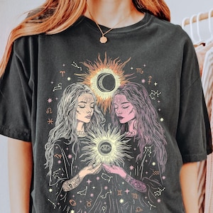 May include: Black oversized t-shirt with a graphic print of two women holding a sun with an eye in the center. The sun is surrounded by zodiac symbols and stars.