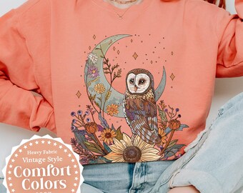 Pode incluir: Camisola cor terracota com um design vintage de uma coruja numa lua crescente com flores. Feita de lã polar macia, com o texto "Comfort Colors". Disponível em versão leve ou pesada. Medidas em cm.