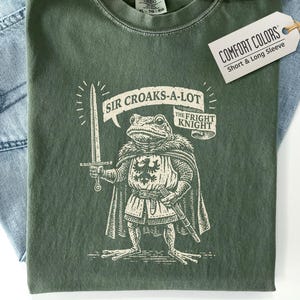 May include: A moss green t-shirt featuring a cream-colored graphic of a frog dressed as a knight, holding a sword. The text reads "SIR CROAKS-A-LOT" and "THE FRIGHT KNIGHT." A white tag attached to the collar displays the text "COMFORT COLORS."