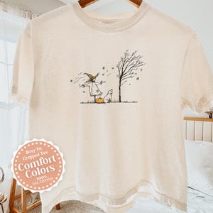 May include: An ivory boxy fit cropped t-shirt featuring a black line art illustration of a scarecrow wearing a hat, a small dog, and a pumpkin standing near a bare tree. The shirt is made of 100% cotton and includes a circular label on the front.