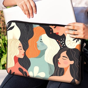 Può includere: Una custodia per laptop con un design astratto colorato che presenta più volti di donne. La custodia è nera con una cerniera bianca ed è tenuta da una persona.