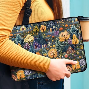 May include: A black laptop sleeve with a colorful autumn-themed design featuring houses, trees, and flowers. The sleeve is held by a person wearing a yellow sweater and blue jeans.